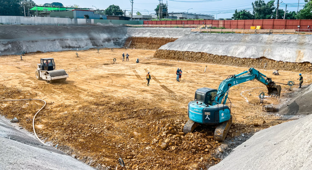 開挖坑四周為斜向放坡而非垂直擋土壁，坡面覆蓋噴凝土保護；坑底已整平並以壓路機壓實，上緣紅色圍籬外即為既有道路與廠房，可看出放坡佔去的腹地範圍
